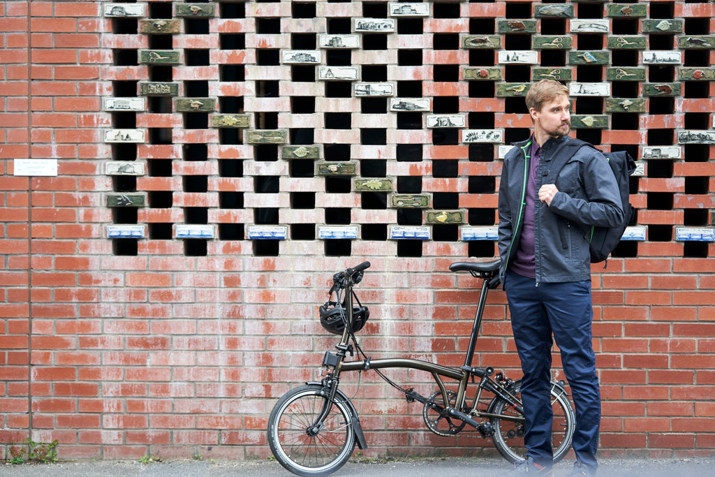 Man standing with a folding bike against a brick wall, wearing Vulpine cycling clothing