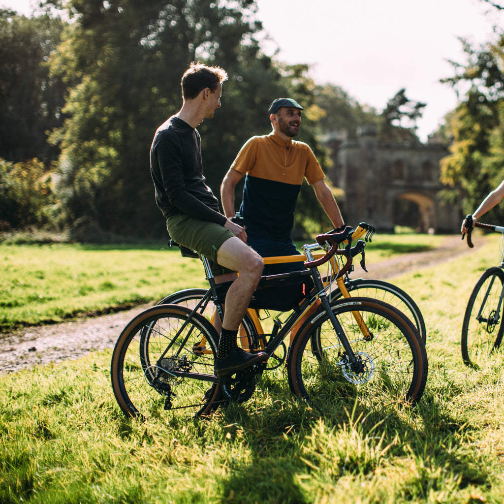 Two cyclists, dressed in casual Vulpine cycling gear, are having a friendly conversation next to their bikes on a grassy path, with an old stone building in the background