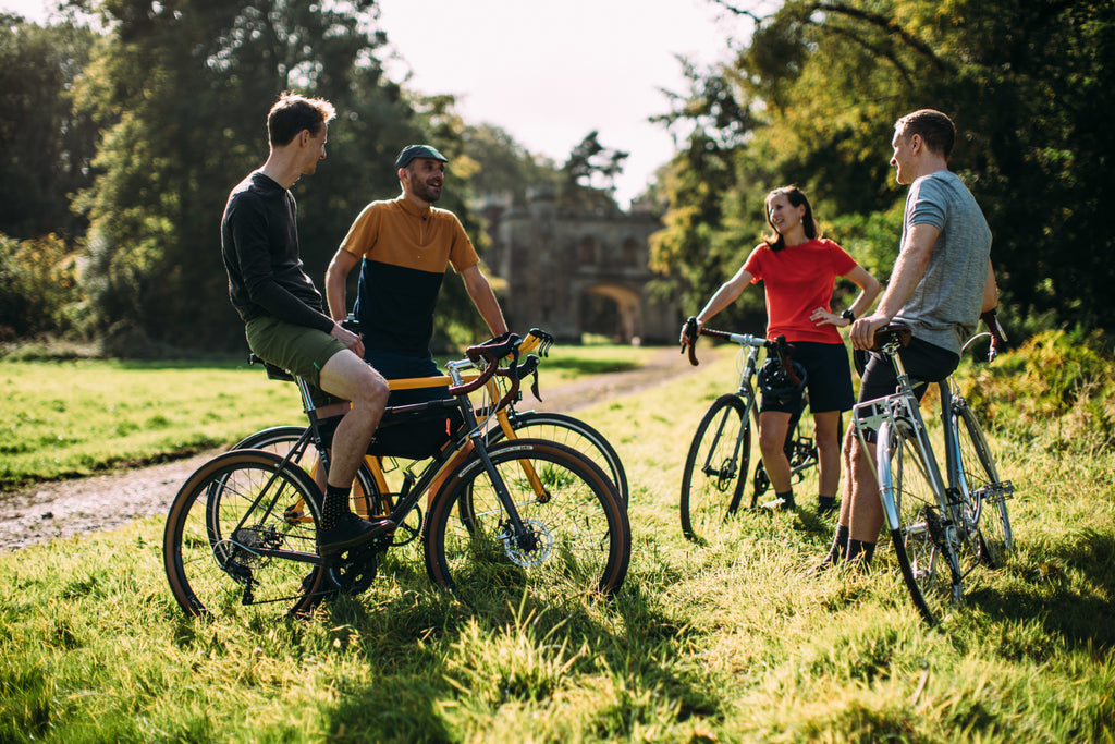 Four cyclists, dressed in casual Vulpine cycling gear, are having a friendly conversation next to their bikes on a grassy path, with an old stone building in the background